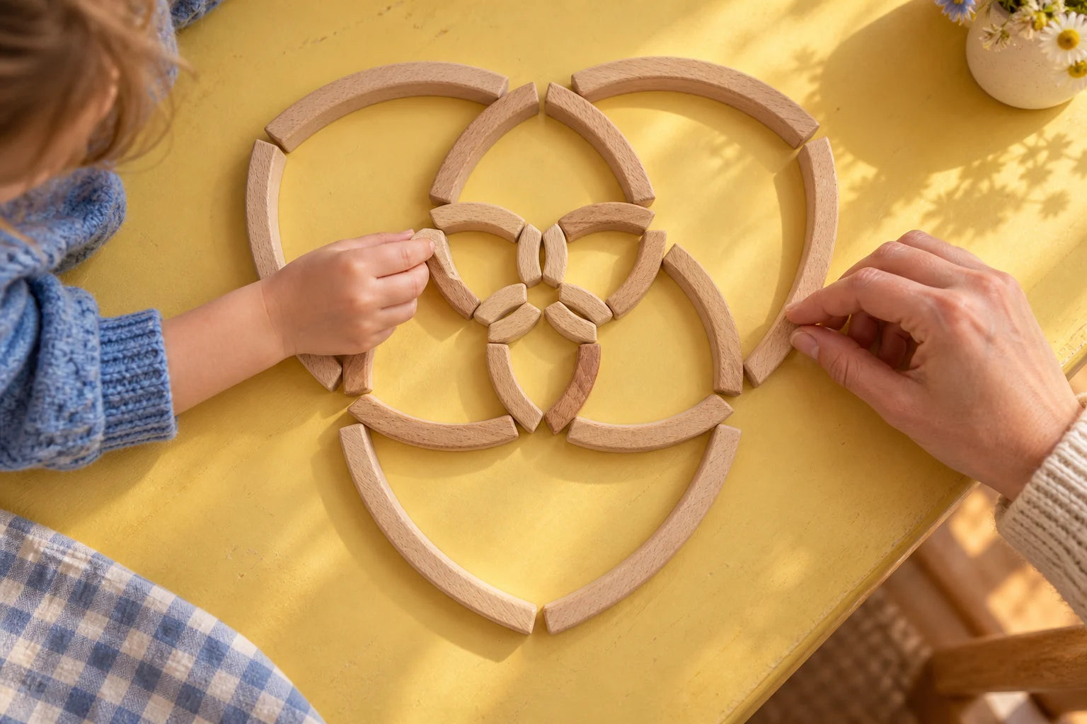 A child and adult arranging graduated curved wooden blocks into a flower on a yellow table