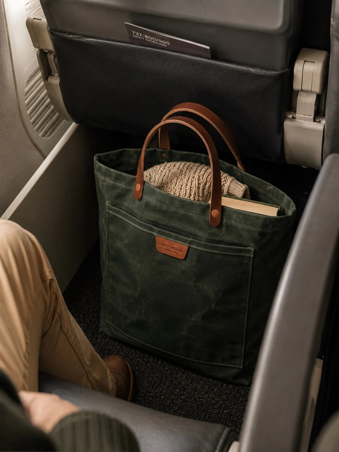 Coal Tote tucked at an airplane seat holding a sweater and a book.