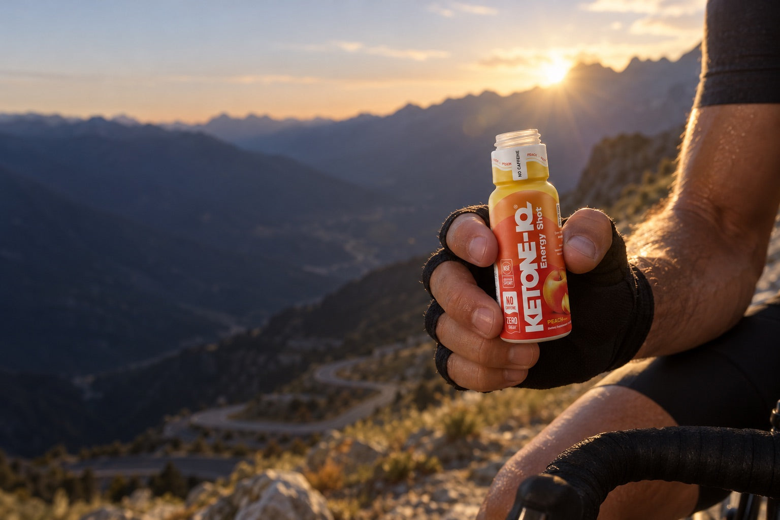 A cyclist's gloved hand holding a Ketone-IQ Peach shot at sunrise on a mountain pass