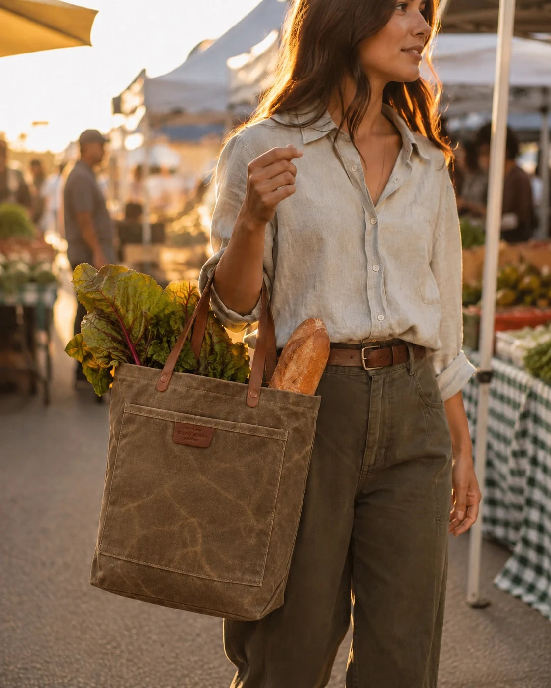 A woman carries the Field Tan Coal Tote, loaded with greens and a baguette, through a sunlit farmers market.