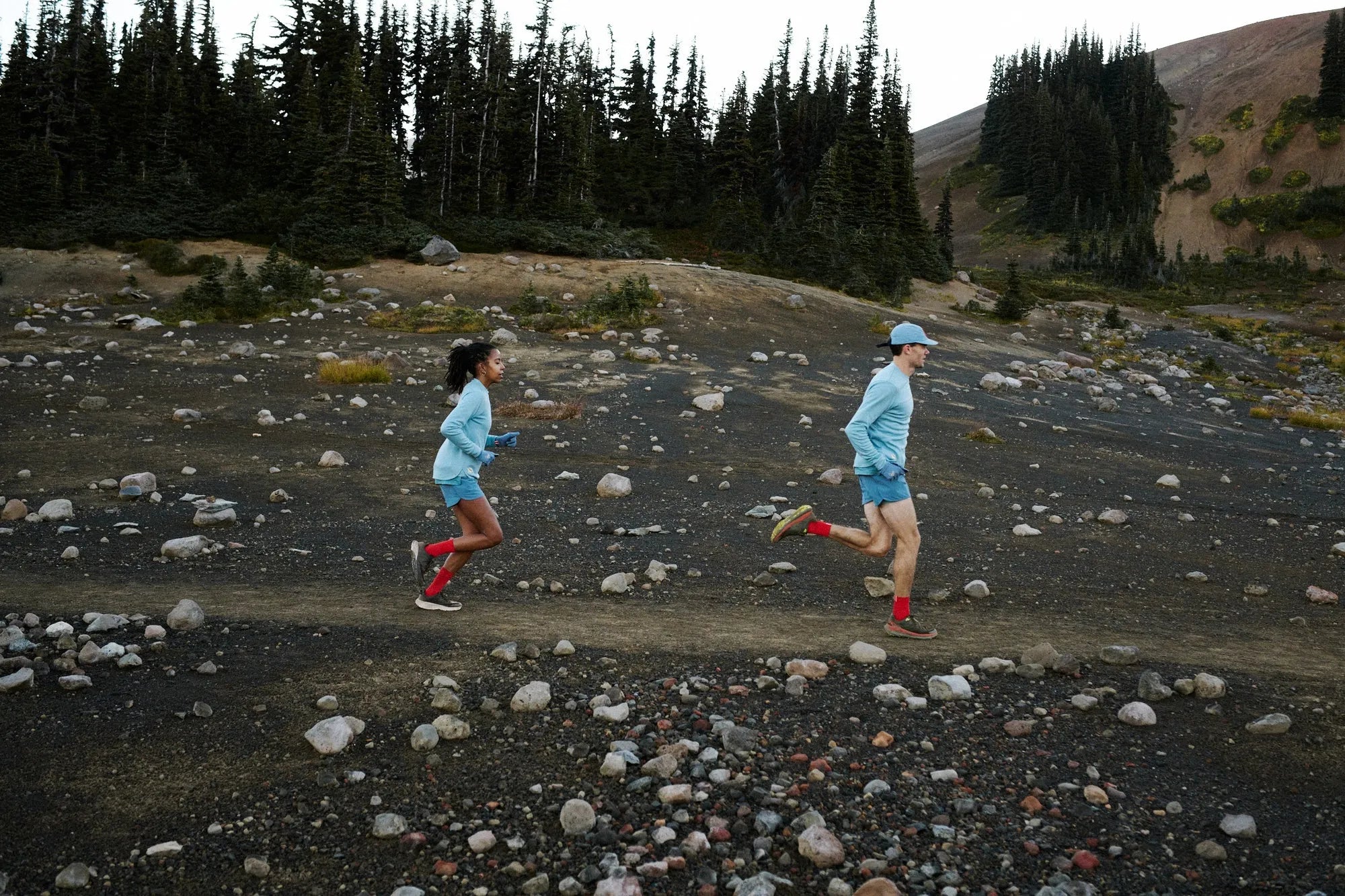 Two runners crossing a rocky trail under an overcast sky