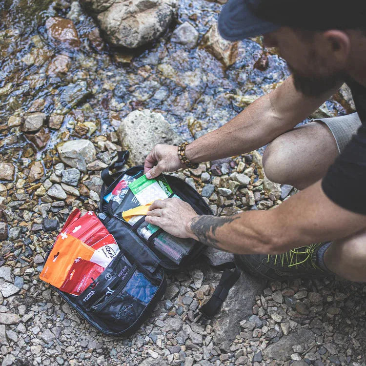 A hiker crouched on a rocky creek bank, reaching into an open MyFAK