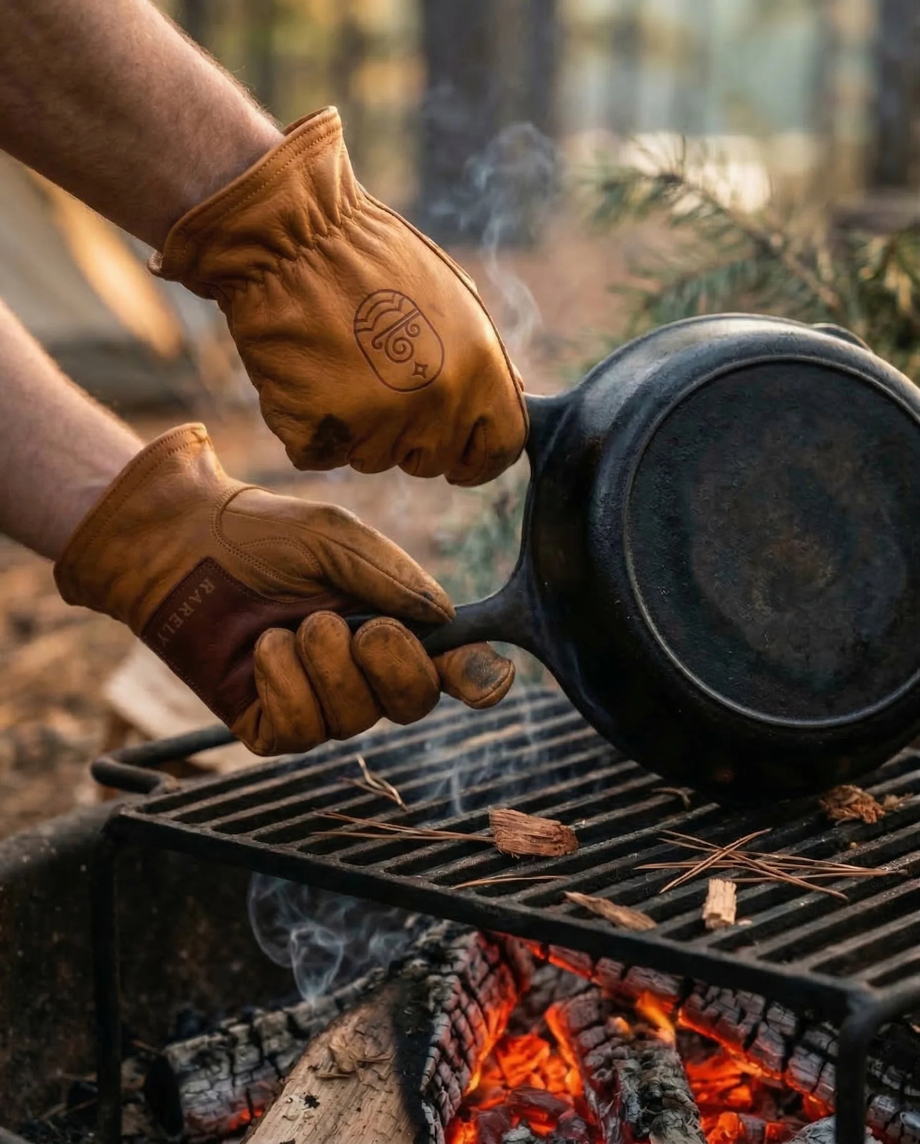 Gloved hands lifting a cast-iron skillet off a campfire grate